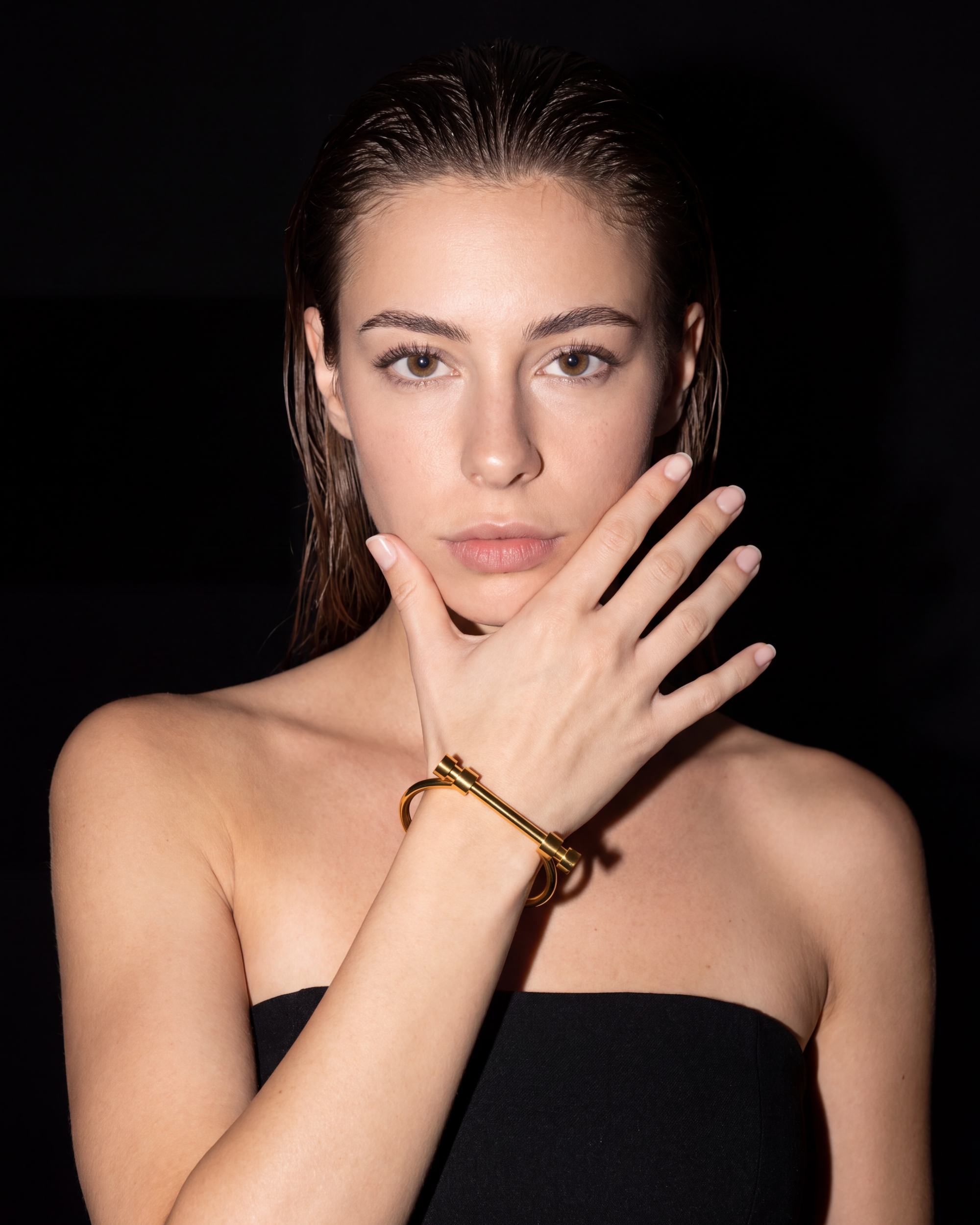 A woman with wet hair posing against a black background, wearing a gold bangle bracelet on her wrist and a black strapless top.