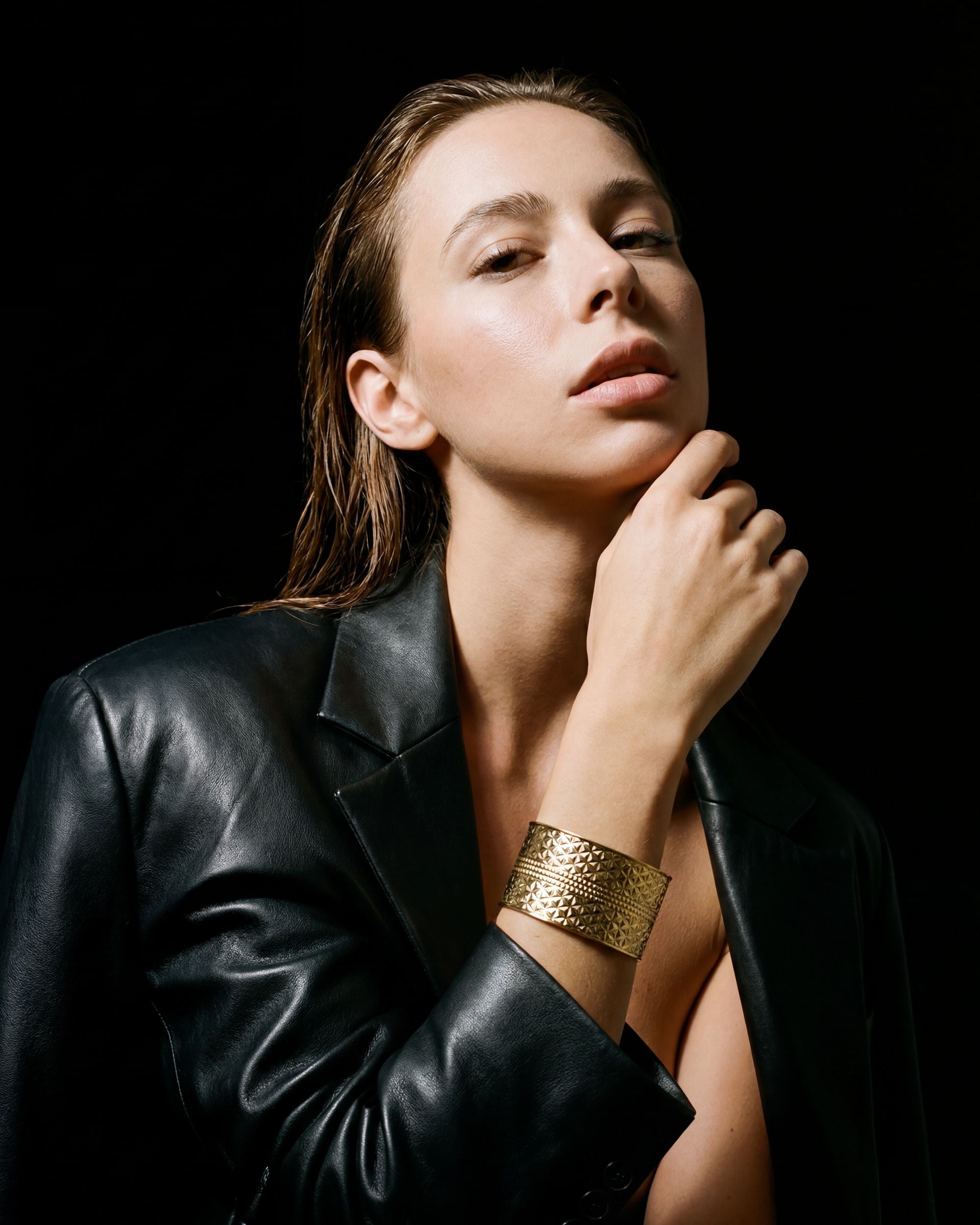 Woman wearing a black leather jacket and gold bracelet against a dark background